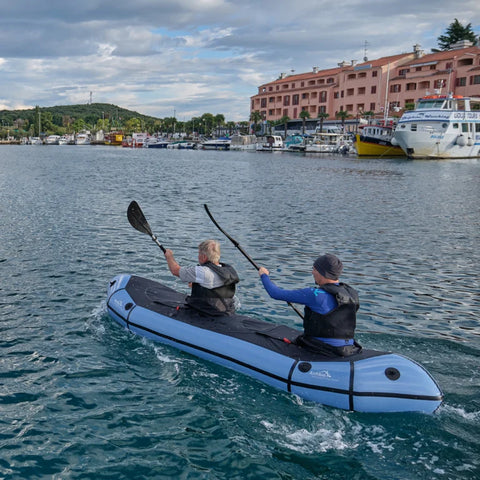 Lyseblå Anfibio Rebel Duo packraft med spruttrekk padles på åpent hav ved en marina. En lett og rask tomanns packraft for tur og trening. Foto: Lisa Kristin Schröter.