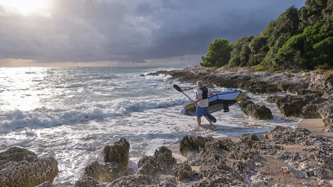 En person bærer en Anfibio Revo CL packraft ned til vannet på en steinstrand i Kroatia. Viser hvor lett båten er å transportere selv for én person. Foto: Anfibio Packrafting.