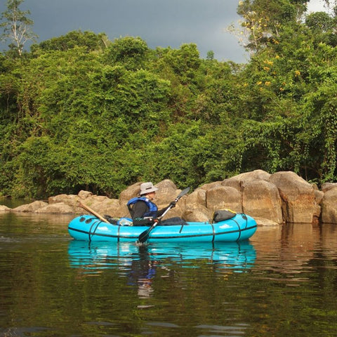 Blå Anfibio Sigma TXL packraft på rolig elv med tett jungelvegetasjon og steiner i bakgrunnen. Viser båtens stabilitet på ekspedisjon. Foto: Anfibio Packrafting.