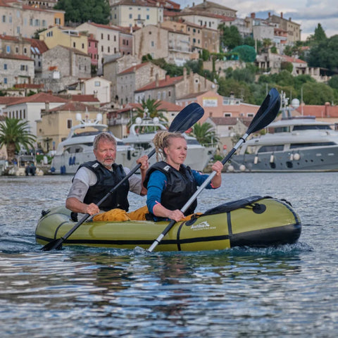o personer padler en grønn Anfibio Sigma TXL packraft i en havn med historisk bebyggelse i bakgrunnen. Lett tomanns packraft for par. Foto: Anfibio Packrafting.